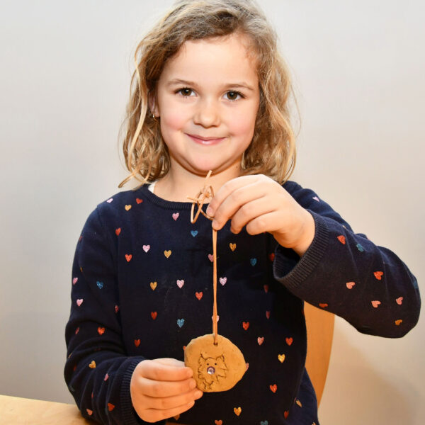 Ein junges Mädchen mit hellbraunem Haar hält ein rundes, verziertes Handwerk aus Ton an einer Schnur hoch. Sie trägt einen marineblauen Pullover mit bunten Herzmustern.
