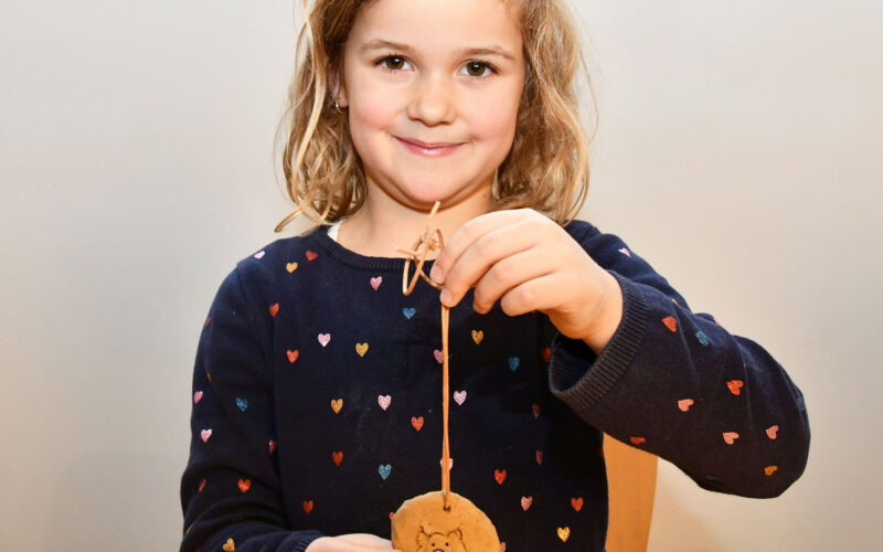 Ein junges Mädchen mit hellbraunem Haar hält ein rundes, verziertes Handwerk aus Ton an einer Schnur hoch. Sie trägt einen marineblauen Pullover mit bunten Herzmustern.