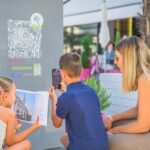Zwei Kinder und eine Frau interagieren mit einem Informationsdisplay im Freien; ein Kind scannt einen QR-Code, während das andere eine Broschüre hält.