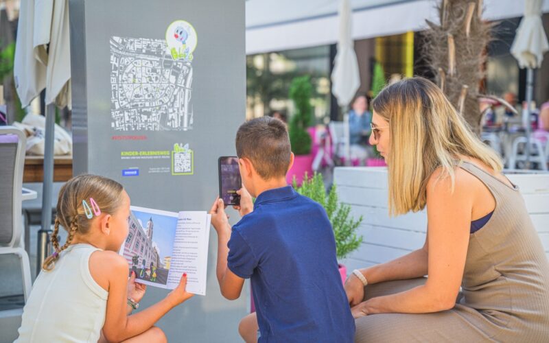 Zwei Kinder und eine Frau interagieren mit einem Informationsdisplay im Freien; ein Kind scannt einen QR-Code, während das andere eine Broschüre hält.
