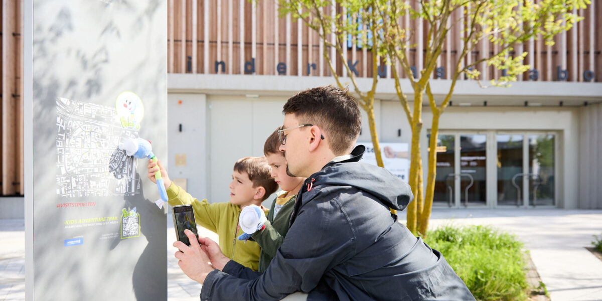 Ein Erwachsener und zwei Kinder verwenden im Freien ein Kartendisplay; der Erwachsene hält ein Smartphone, während die Kinder mit blumenförmigen Stöcken zeigen.