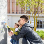 Ein Erwachsener und zwei Kinder verwenden im Freien ein Kartendisplay; der Erwachsene hält ein Smartphone, während die Kinder mit blumenförmigen Stöcken zeigen.