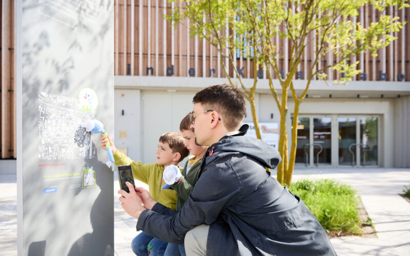 Ein Erwachsener und zwei Kinder verwenden im Freien ein Kartendisplay; der Erwachsene hält ein Smartphone, während die Kinder mit blumenförmigen Stöcken zeigen.