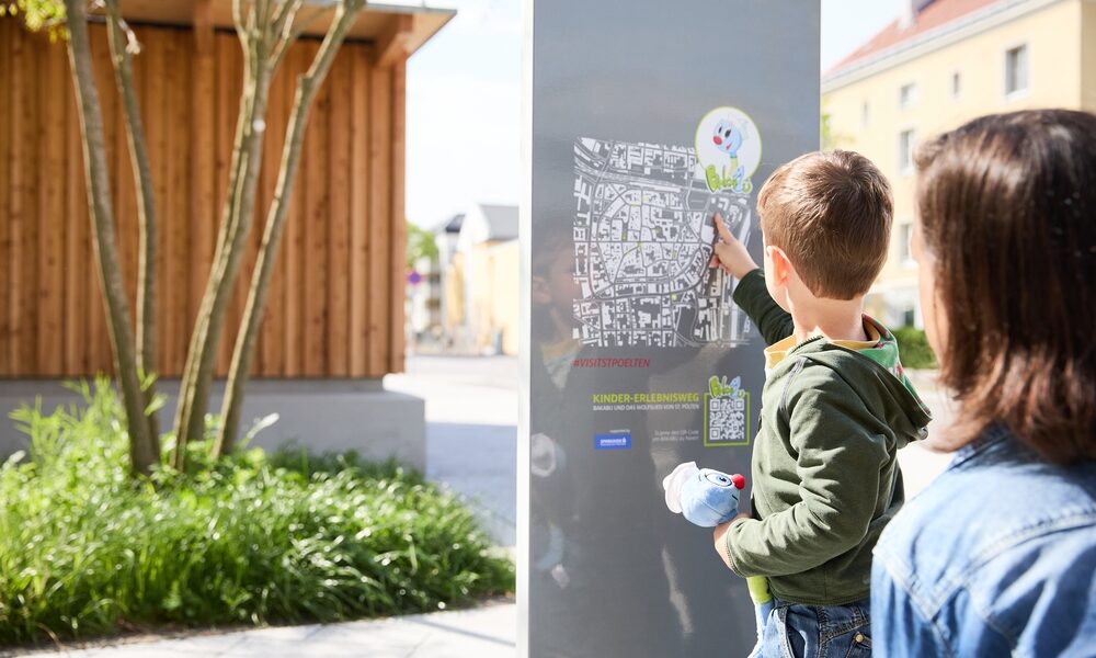 Ein kleiner Junge zeigt auf eine Karte auf einer Informationstafel im Freien, während ein Erwachsener neben ihm steht.