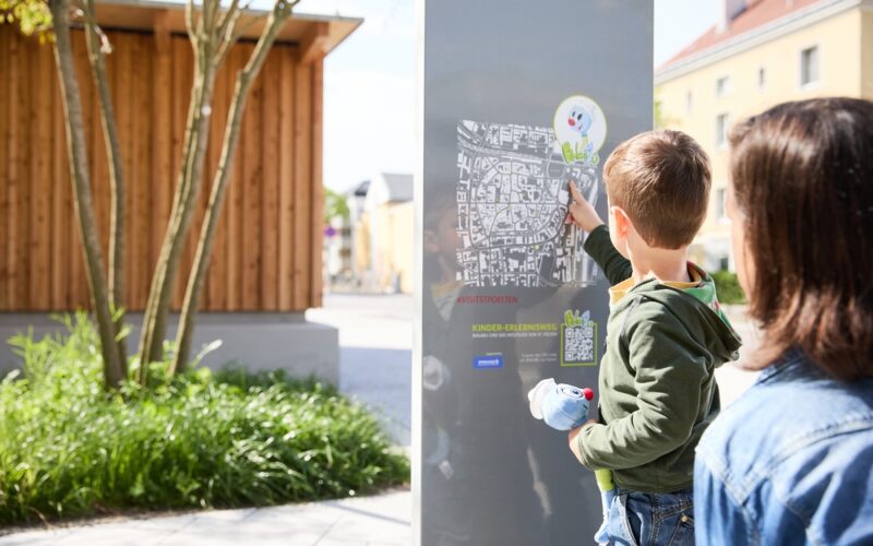 Ein kleiner Junge zeigt auf eine Karte auf einer Informationstafel im Freien, während ein Erwachsener neben ihm steht.