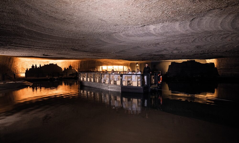 Ein Boot mit flachem Boden und Passagieren fährt durch eine beleuchtete unterirdische Höhle mit strukturierten Steindecken, die sich auf dem stillen Wasser spiegeln.