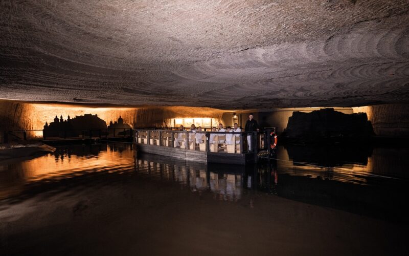 Ein Boot mit flachem Boden und Passagieren fährt durch eine beleuchtete unterirdische Höhle mit strukturierten Steindecken, die sich auf dem stillen Wasser spiegeln.