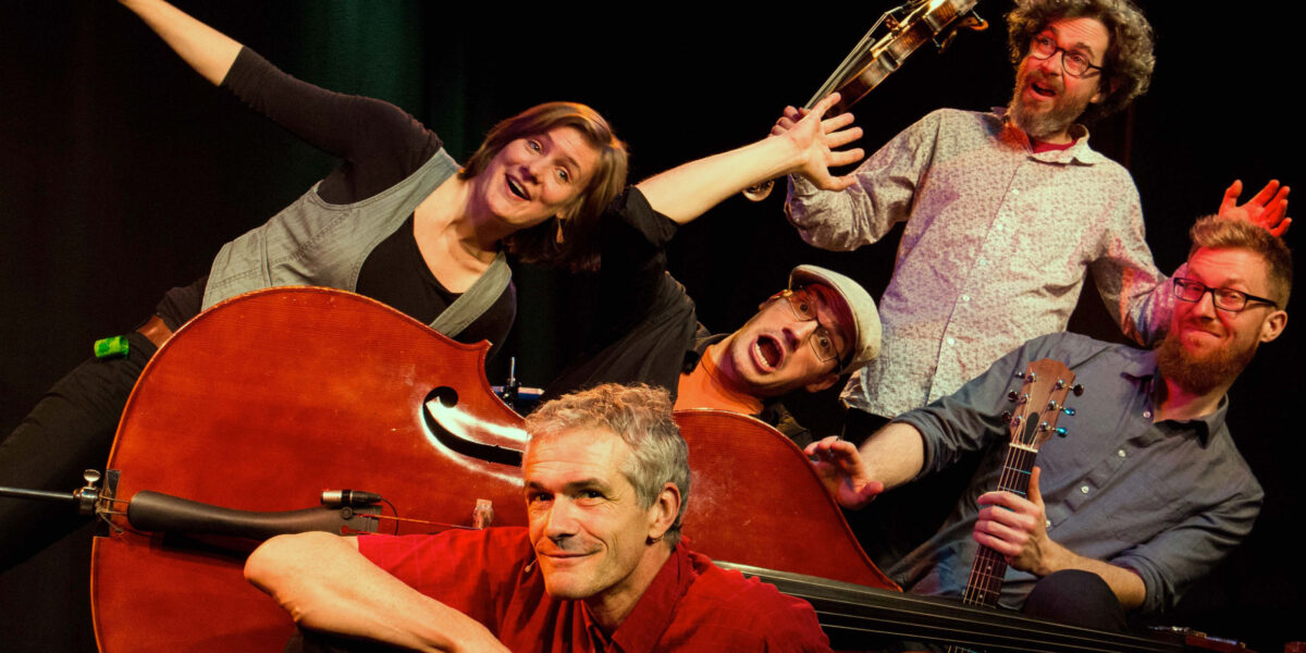 Fünf Erwachsene posieren spielerisch mit Musikinstrumenten, darunter ein Kontrabass, eine Gitarre und eine Geige, vor einem dunklen Hintergrund.