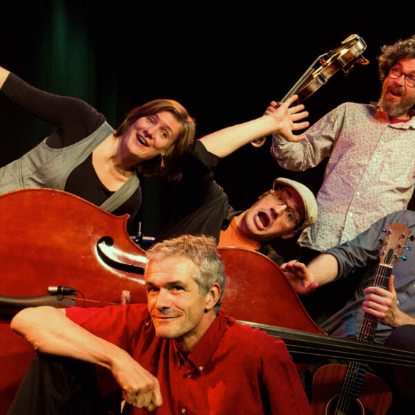 Fünf Erwachsene posieren spielerisch mit Musikinstrumenten, darunter ein Kontrabass, eine Gitarre und eine Geige, vor einem dunklen Hintergrund.