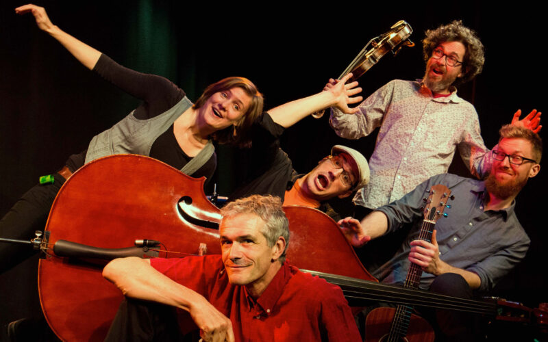 Fünf Erwachsene posieren spielerisch mit Musikinstrumenten, darunter ein Kontrabass, eine Gitarre und eine Geige, vor einem dunklen Hintergrund.