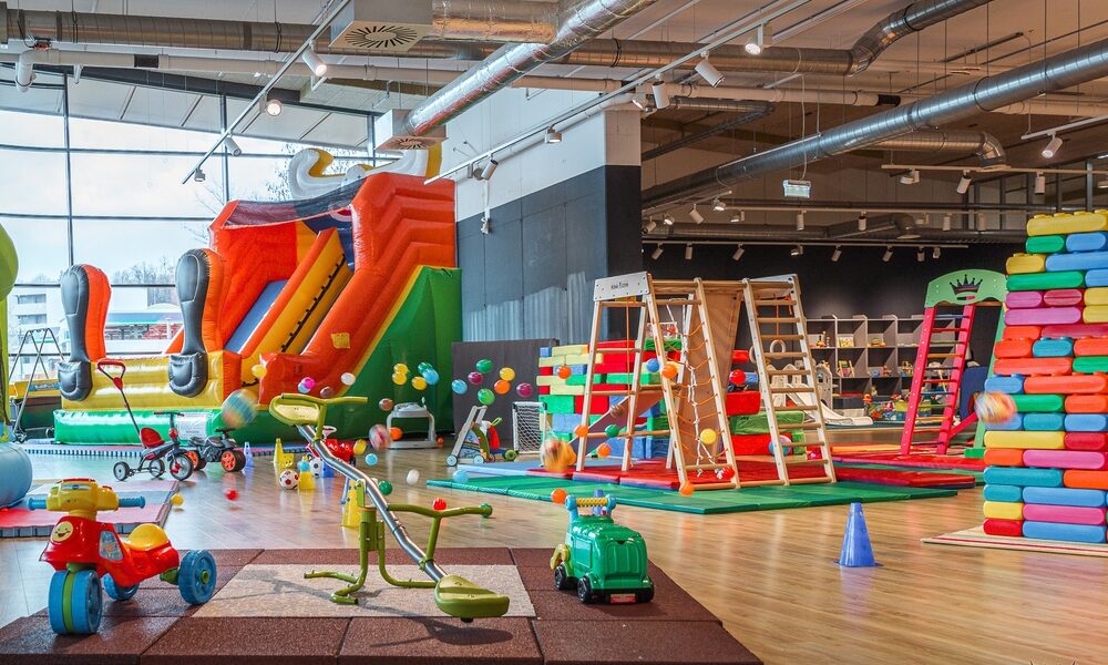 Indoor-Kinderspielbereich mit einer großen aufblasbaren Rutsche, Klettergerüsten, Aufsitzspielzeug, weichen Spielgeräten und farbenfroher Dekoration auf einem Holzboden.
