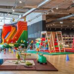 Indoor-Kinderspielbereich mit einer großen aufblasbaren Rutsche, Klettergerüsten, Aufsitzspielzeug, weichen Spielgeräten und farbenfroher Dekoration auf einem Holzboden.