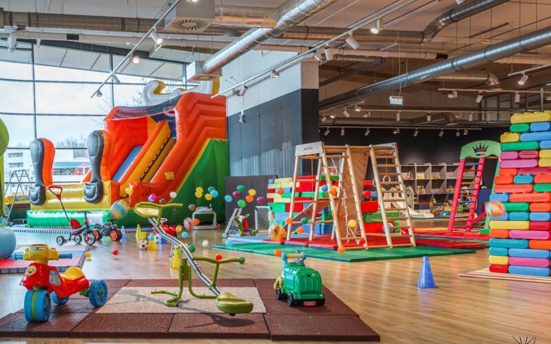 Indoor-Kinderspielbereich mit einer großen aufblasbaren Rutsche, Klettergerüsten, Aufsitzspielzeug, weichen Spielgeräten und farbenfroher Dekoration auf einem Holzboden.