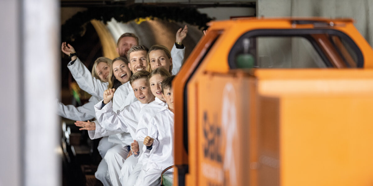 Eine Gruppe von Personen in weißen Overalls sitzt in einem kleinen Zug, lächelt und zeigt nach vorne in einen geschlossenen Tunnelraum. Im Vordergrund ist ein orangefarbenes Fahrzeug zu sehen.
