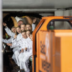 Eine Gruppe von Personen in weißen Overalls sitzt in einem kleinen Zug, lächelt und zeigt nach vorne in einen geschlossenen Tunnelraum. Im Vordergrund ist ein orangefarbenes Fahrzeug zu sehen.