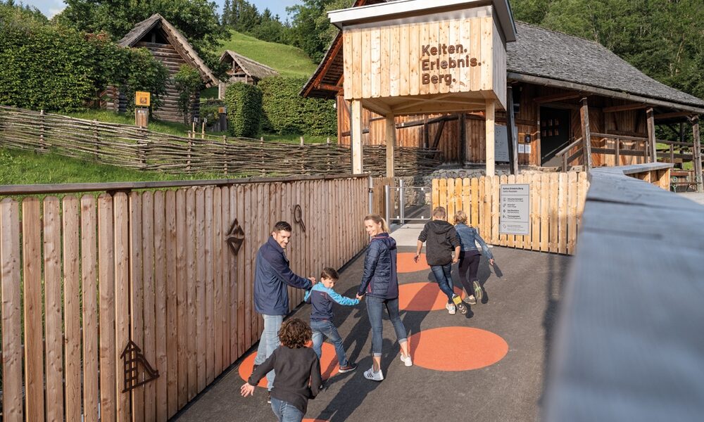 Eine Gruppe von Kindern und Erwachsenen läuft und spielt auf einem Weg mit orangefarbenen Kreisen vor einem Holzgebäude mit der Aufschrift "Kelten Erlebnis Berg" in einer ländlichen, hügeligen Gegend.