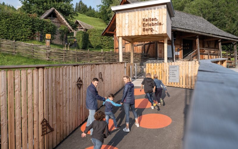 Eine Gruppe von Kindern und Erwachsenen läuft und spielt auf einem Weg mit orangefarbenen Kreisen vor einem Holzgebäude mit der Aufschrift "Kelten Erlebnis Berg" in einer ländlichen, hügeligen Gegend.