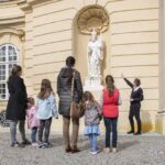 Eine Gruppe von Kindern und Erwachsenen hört einem Führer zu, der eine weiße Statue an der Außenwand eines historischen Gebäudes erklärt.