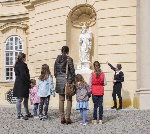 Eine Gruppe von Kindern und Erwachsenen hört einem Führer zu, der eine weiße Statue an der Außenwand eines historischen Gebäudes erklärt.