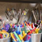 Auf einem Regal sind verschiedene Pinsel und Behälter mit Markern, Stiften, Scheren und anderem Kunstzubehör angeordnet.