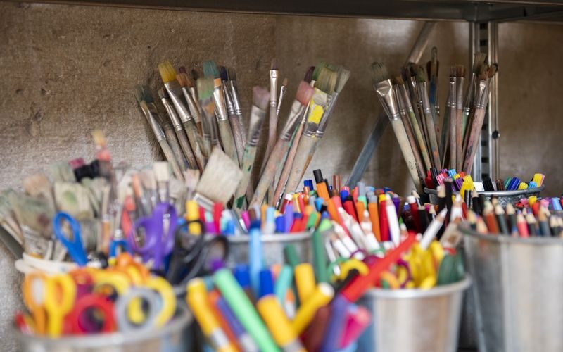 Auf einem Regal sind verschiedene Pinsel und Behälter mit Markern, Stiften, Scheren und anderem Kunstzubehör angeordnet.
