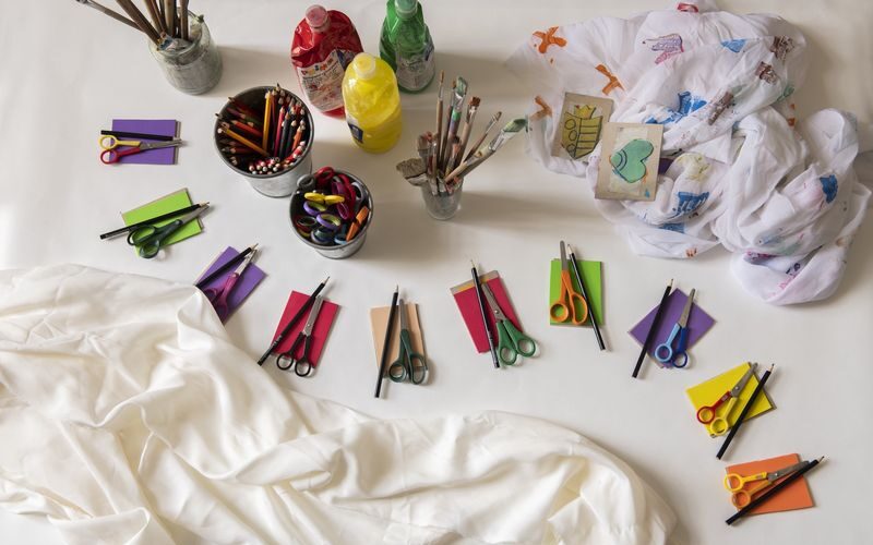 Kunstzubehör wie Scheren, Buntstifte, Farbflaschen, Pinsel, farbiges Papier und Stoff sind ordentlich auf einer weißen Fläche angeordnet.