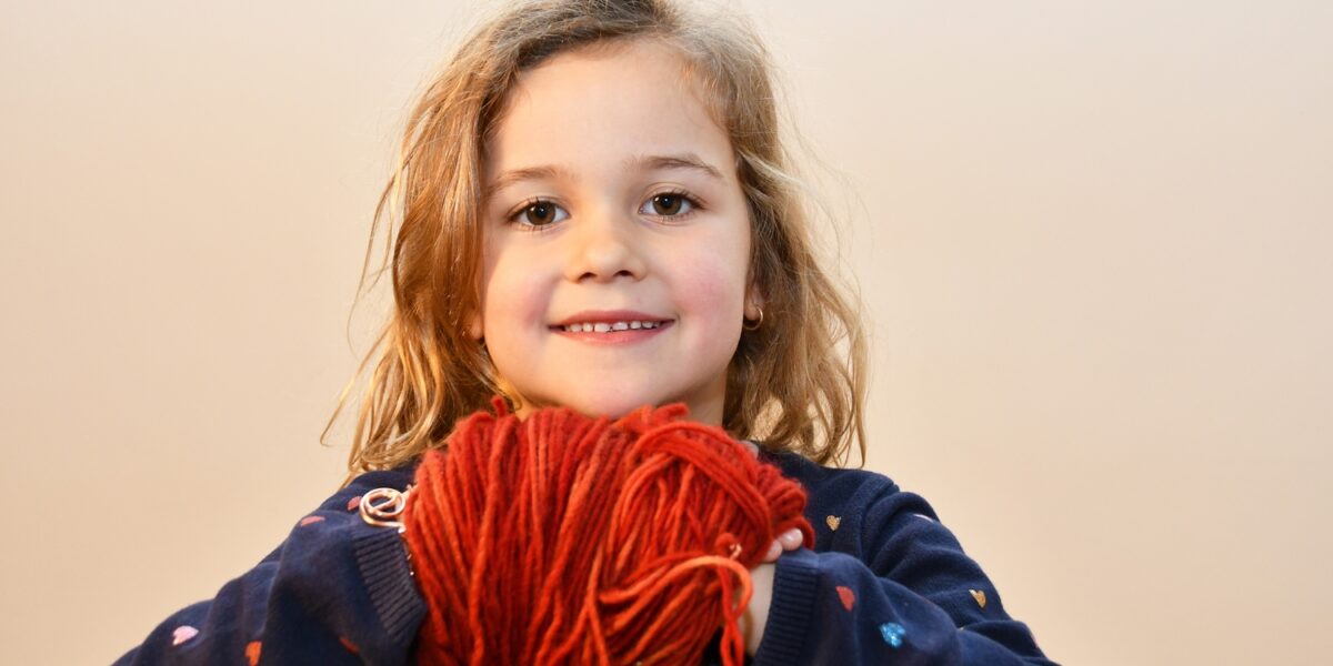 Ein junges Mädchen mit gewelltem blondem Haar lächelt und hält ein großes rotes Garnknäuel in beiden Händen. Sie trägt einen dunklen Pullover mit kleinen bunten Herzmustern.