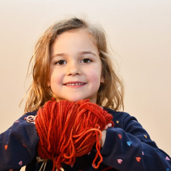 Ein junges Mädchen mit gewelltem blondem Haar lächelt und hält ein großes rotes Garnknäuel in beiden Händen. Sie trägt einen dunklen Pullover mit kleinen bunten Herzmustern.