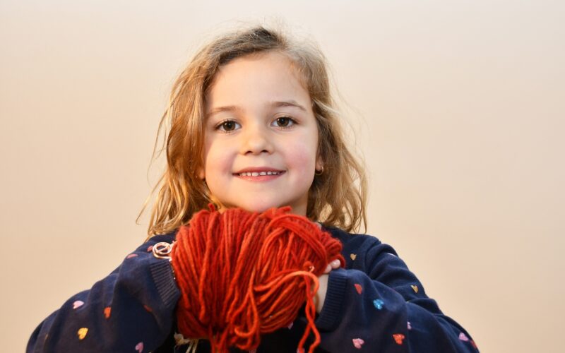 Ein junges Mädchen mit gewelltem blondem Haar lächelt und hält ein großes rotes Garnknäuel in beiden Händen. Sie trägt einen dunklen Pullover mit kleinen bunten Herzmustern.