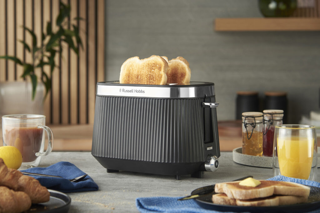 Ein schwarzer Russell Hobbs-Toaster mit zwei Scheiben getoastetem Brot steht auf einem für das Frühstück gedeckten Küchentisch mit Saft, Kaffee, Gebäck und Marmeladengläsern.