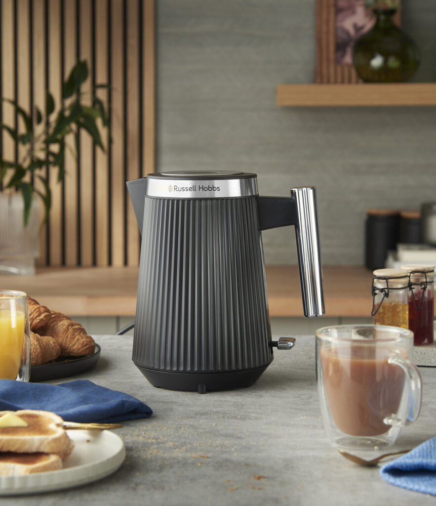 Ein schwarzer Russell Hobbs-Wasserkocher steht auf dem Küchentisch neben einer Tasse Tee, Toast, Orangensaft, Croissants und Gläsern mit Konserven.