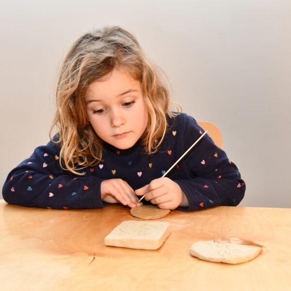 Ein kleines Kind sitzt an einem Tisch und schnitzt mit einem Metallwerkzeug Motive in ein Stück Ton, während auf dem Tisch zwei Tonplatten liegen.