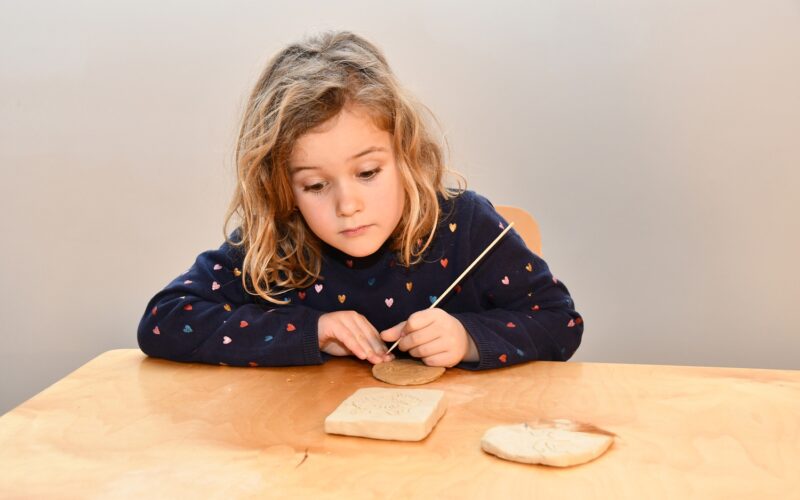 Ein kleines Kind sitzt an einem Tisch und schnitzt mit einem Metallwerkzeug Motive in ein Stück Ton, während auf dem Tisch zwei Tonplatten liegen.