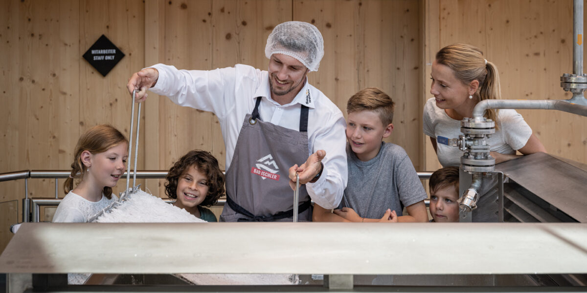 Ein Mann mit Haarnetz und Schürze rührt einen großen Bottich mit Quark, während vier Kinder und eine Frau in einem hölzernen Raum aufmerksam zusehen.