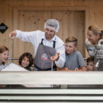 Ein Mann mit Haarnetz und Schürze rührt einen großen Bottich mit Quark, während vier Kinder und eine Frau in einem hölzernen Raum aufmerksam zusehen.
