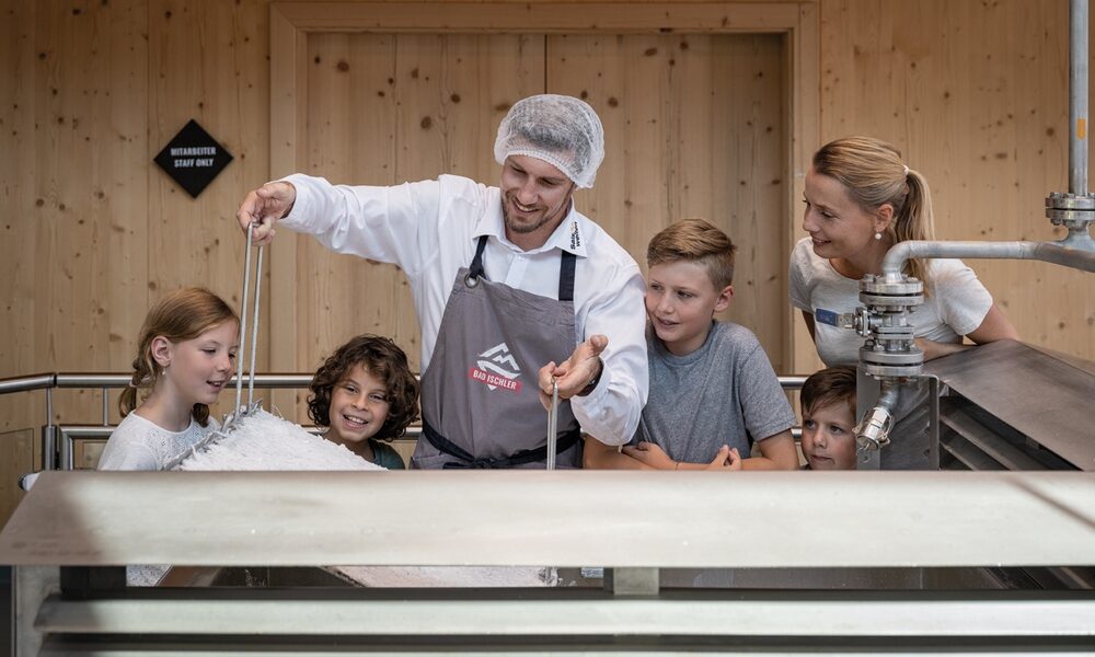 Ein Mann mit Schürze und Haarnetz zeigt vier Kindern und einer Frau in einem hölzernen Raum, wie man Käse herstellt.