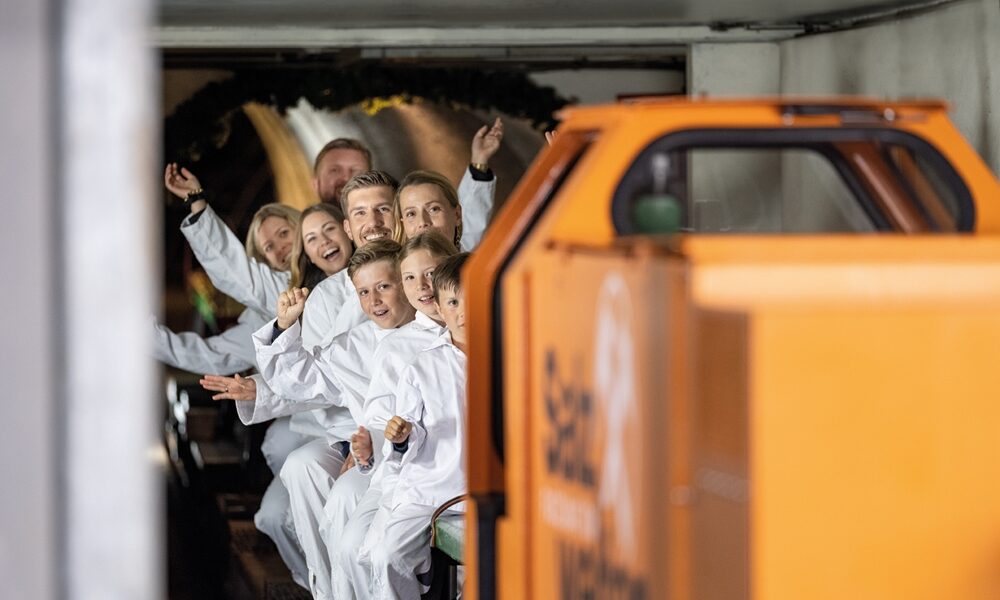 Eine Gruppe von Menschen in weißen Overalls sitzt in einem kleinen U-Bahn-Zug, lächelt und zeigt nach vorne, während sie durch einen Tunnel fahren.