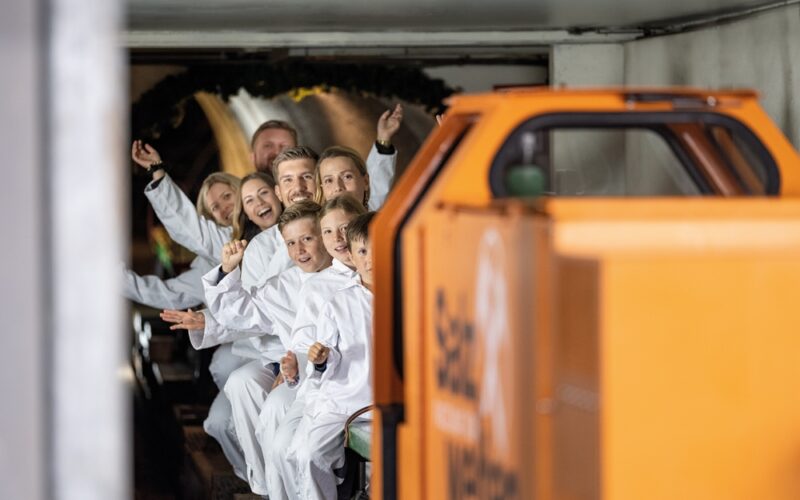 Eine Gruppe von Menschen in weißen Overalls sitzt in einem kleinen U-Bahn-Zug, lächelt und zeigt nach vorne, während sie durch einen Tunnel fahren.