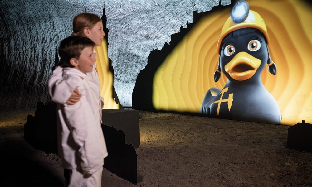 Zwei Kinder in weißer Kleidung betrachten eine große beleuchtete Anzeige einer Cartoon-Ente, die einen Bergmannshelm trägt, in einer unterirdischen Höhle oder einem Tunnel.