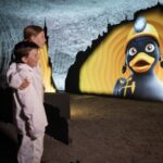 Zwei Kinder in weißer Kleidung betrachten eine große beleuchtete Anzeige einer Cartoon-Ente, die einen Bergmannshelm trägt, in einer unterirdischen Höhle oder einem Tunnel.