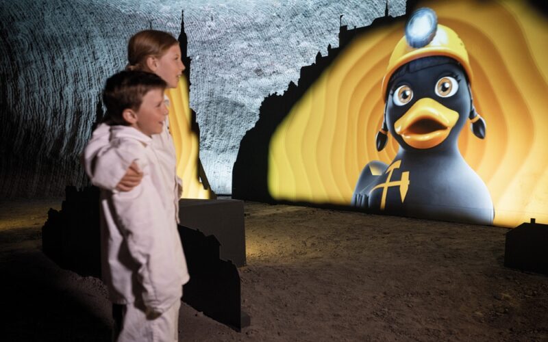 Zwei Kinder in weißer Kleidung betrachten eine große beleuchtete Anzeige einer Cartoon-Ente, die einen Bergmannshelm trägt, in einer unterirdischen Höhle oder einem Tunnel.
