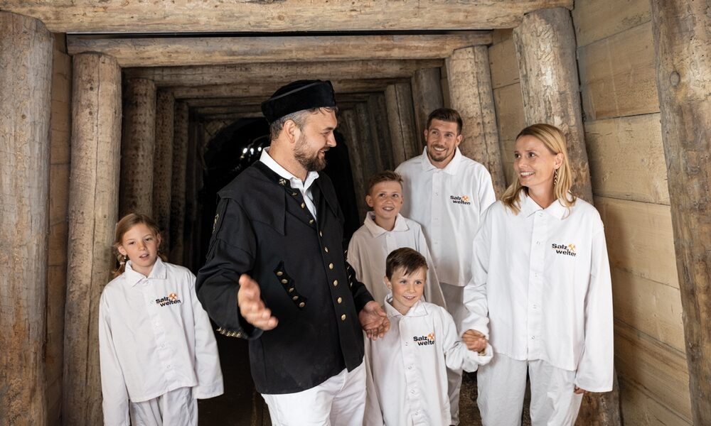 Ein Mann in schwarzer Uniform führt eine Gruppe von Kindern und Erwachsenen in weißer Kleidung durch einen hölzernen Minenstollen.