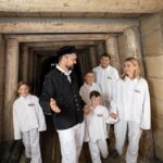 Ein Mann in schwarzer Uniform führt eine Gruppe von Kindern und Erwachsenen in weißer Kleidung durch einen hölzernen Minenstollen.