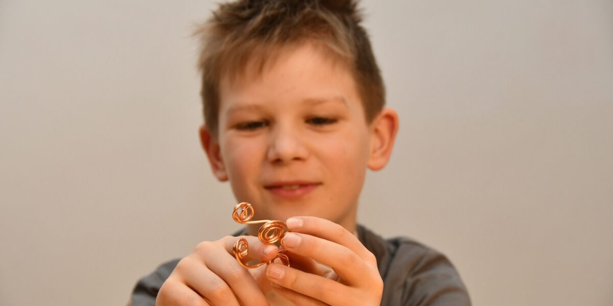 Ein Junge mit unordentlichem Haar hält und betrachtet eine kleine Drahtskulptur mit Spiralformen und konzentriert sich auf das Objekt in seinen Händen.