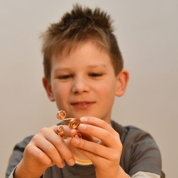 Ein Junge mit unordentlichem Haar hält und betrachtet eine kleine Drahtskulptur mit Spiralformen und konzentriert sich auf das Objekt in seinen Händen.