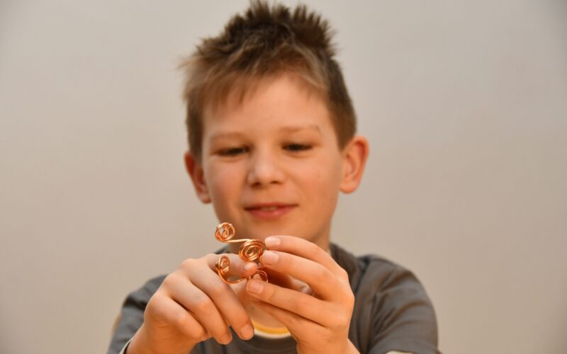 Ein Junge mit unordentlichem Haar hält und betrachtet eine kleine Drahtskulptur mit Spiralformen und konzentriert sich auf das Objekt in seinen Händen.