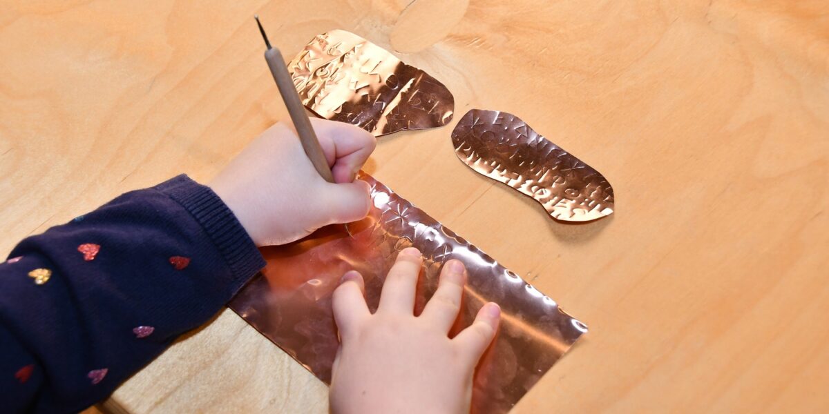 Ein Kind benutzt einen Stift, um ein Motiv auf ein Blatt Metallfolie auf einem Holztisch zu ätzen, wobei zwei abgeschnittene Folienstücke daneben liegen.