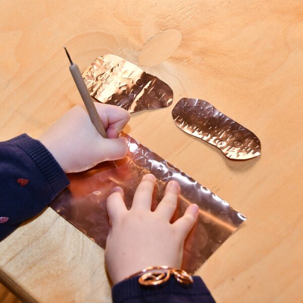 Ein Kind benutzt einen Stift, um ein Motiv auf ein Blatt Metallfolie auf einem Holztisch zu ätzen, wobei zwei abgeschnittene Folienstücke daneben liegen.