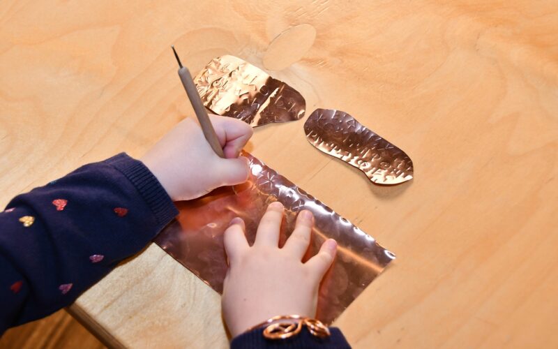 Ein Kind benutzt einen Stift, um ein Motiv auf ein Blatt Metallfolie auf einem Holztisch zu ätzen, wobei zwei abgeschnittene Folienstücke daneben liegen.
