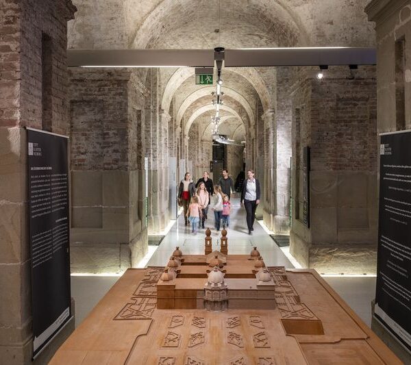 Eine Gruppe von Personen geht durch einen gewölbten Steinflur, in dessen Vordergrund Ausstellungstafeln und ein hölzernes Architekturmodell zu sehen sind.